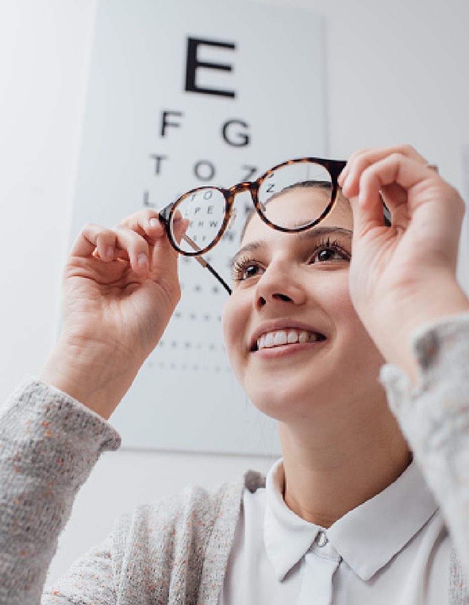 Frau steht vor Sehzeichentafel, hält Ihre Brille vor ihr Gesicht uns sieht hindurch.